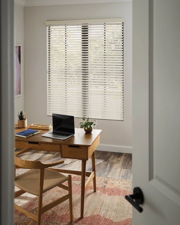 A home office with a wooden desk, chair, and laptop, illuminated by sunlight through blinds.