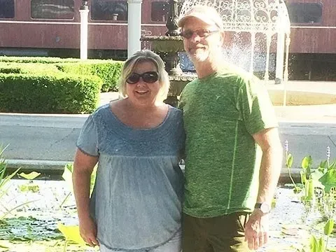 Woman and man standing by a fountain, smiling. The woman wears a blue top; the man, a green shirt and a cap.