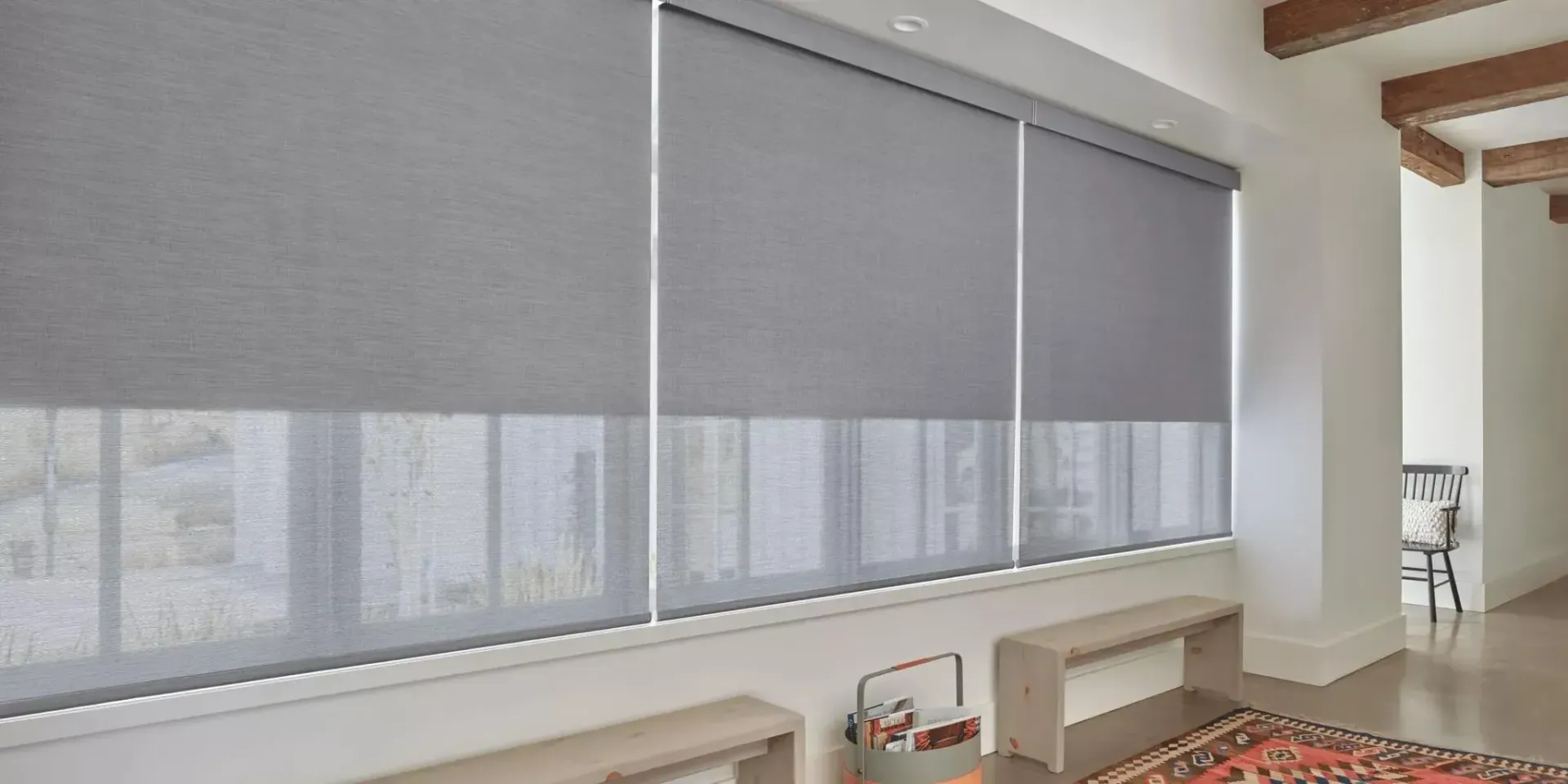 Three gray roller shades partially cover a window, letting in some light. White walls and wooden beams.