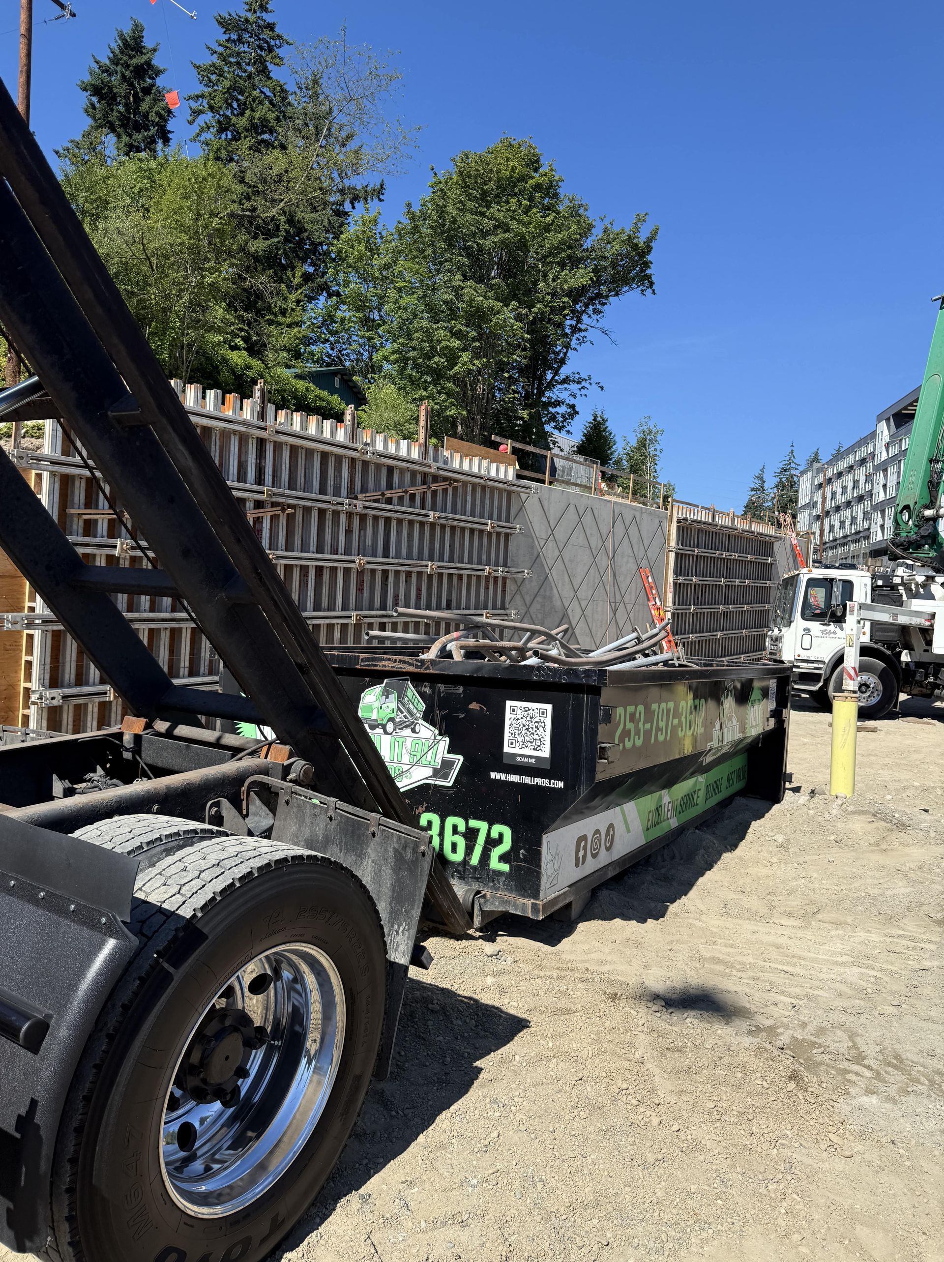Construction site: Dumpster on truck bed next to wooden formwork, trees, and a pump truck.