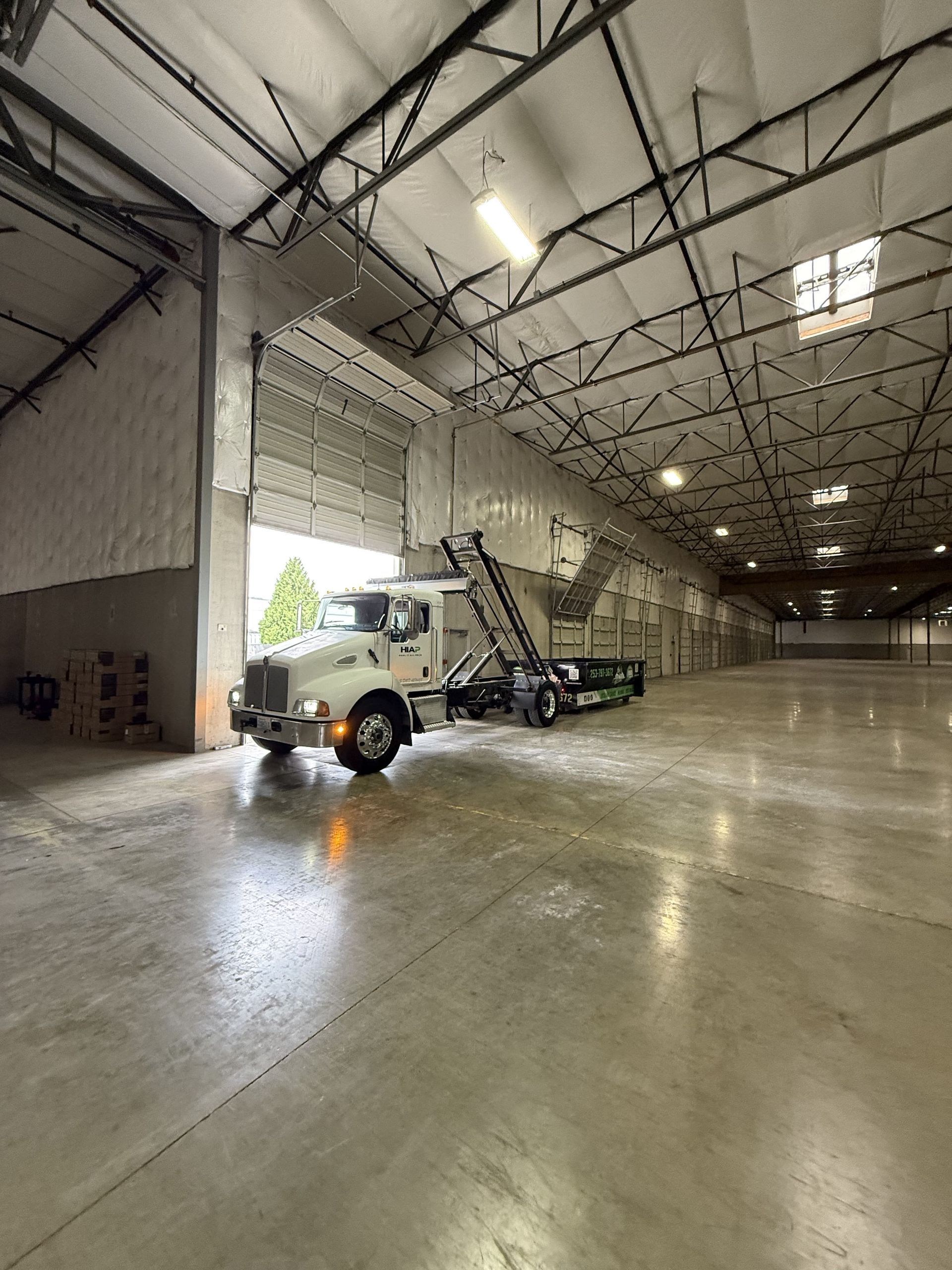 A white truck with a lift inside a large, empty warehouse with an open door.