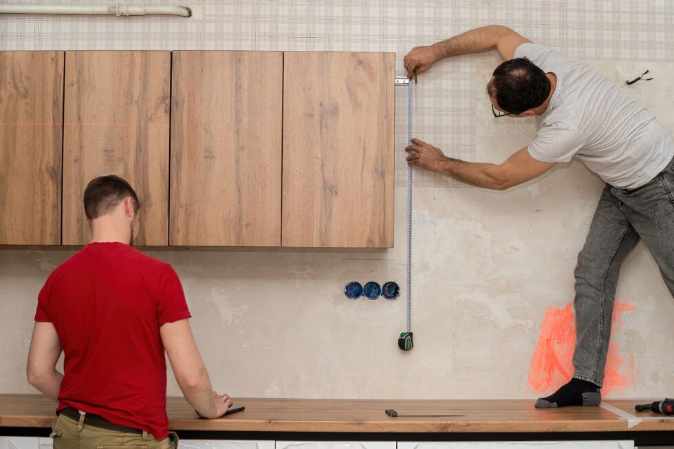 Two men are measuring a cabinet with a tape measure in a kitchen.