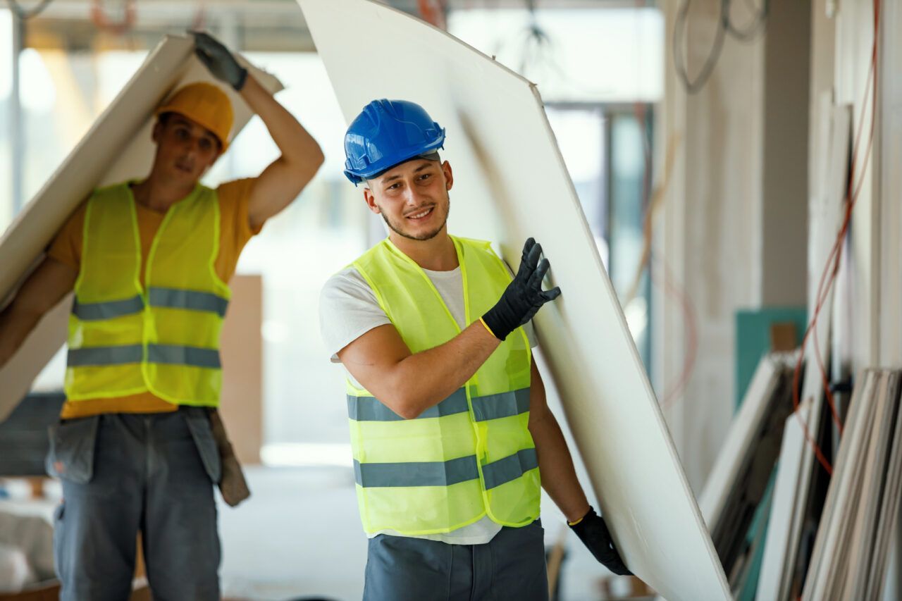 Two construction workers are carrying a large piece of drywall.