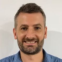 A man with a beard is smiling for the camera while wearing a blue shirt.