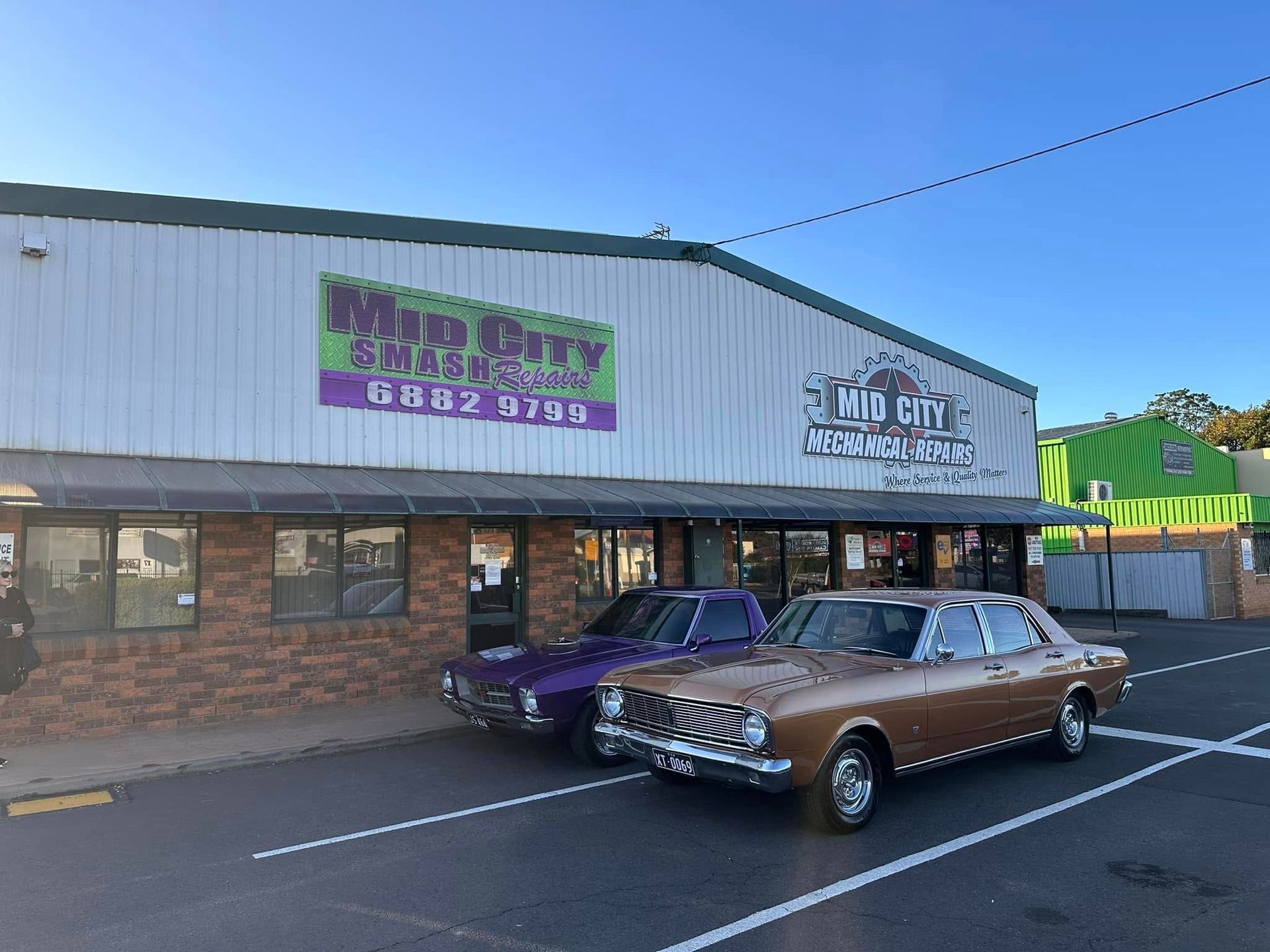 Two classic cars parked in front of Mud City Smash Repairs building with a blue sky background.