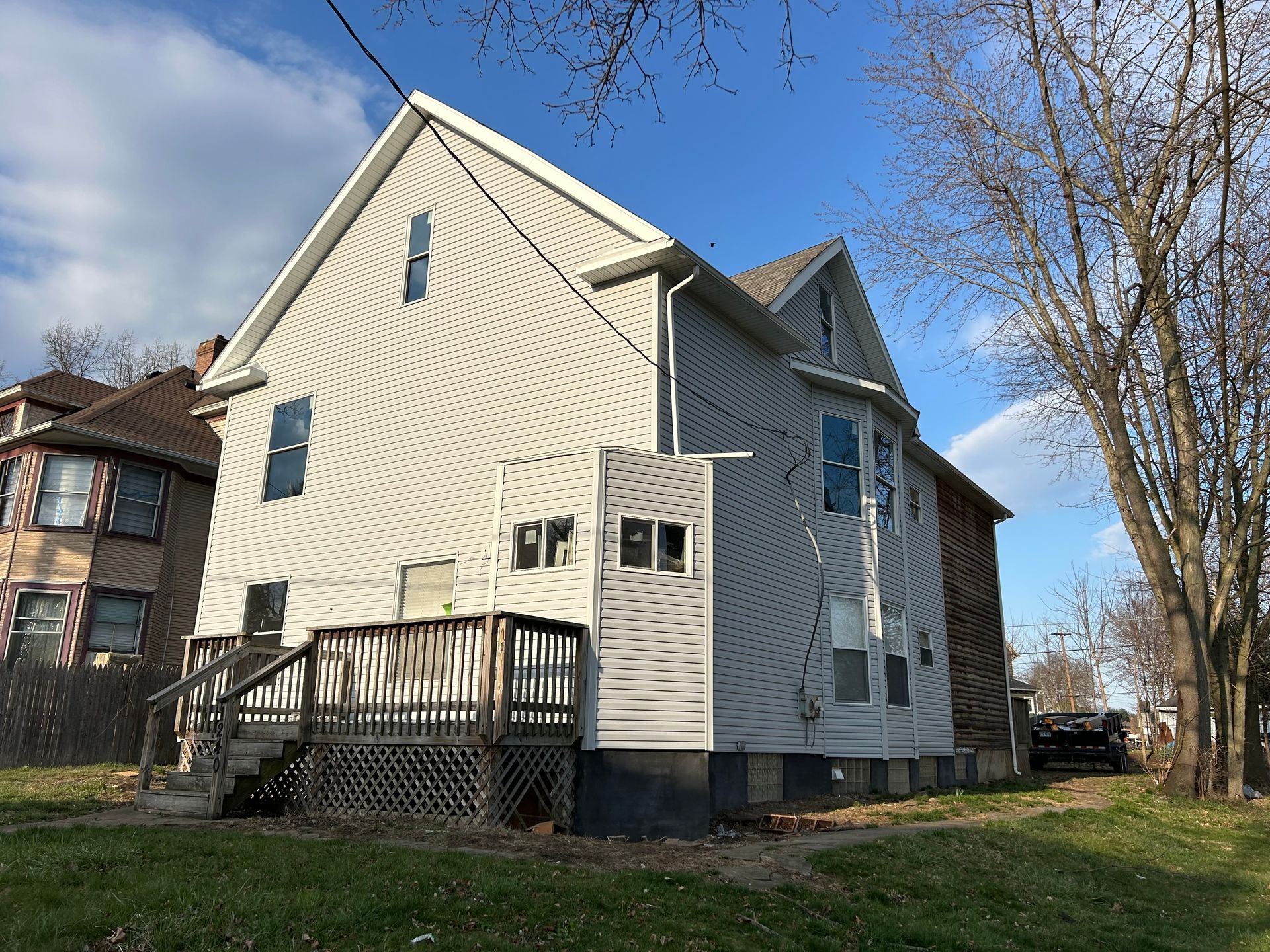Two-story house with light siding and a wooden deck, viewed from the side, with blue sky and bare trees.