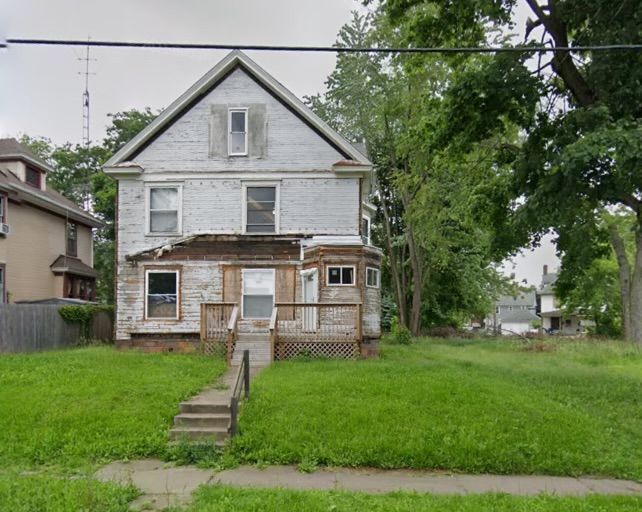 Two-story, weathered white house with porch and overgrown lawn; steps lead to front door.