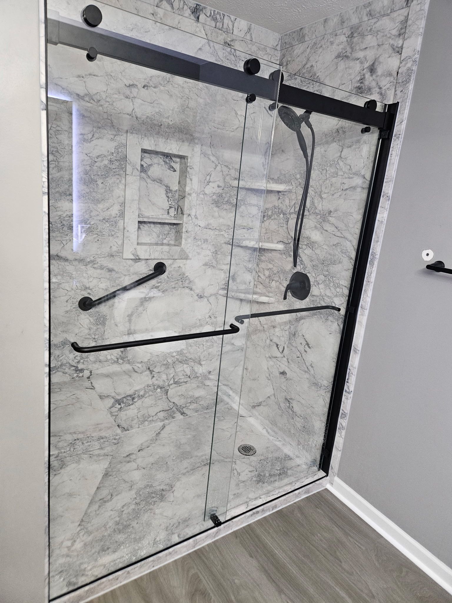 Black-framed glass shower enclosure with gray marble-look walls; features a grab bar, shelves, and a sliding door.