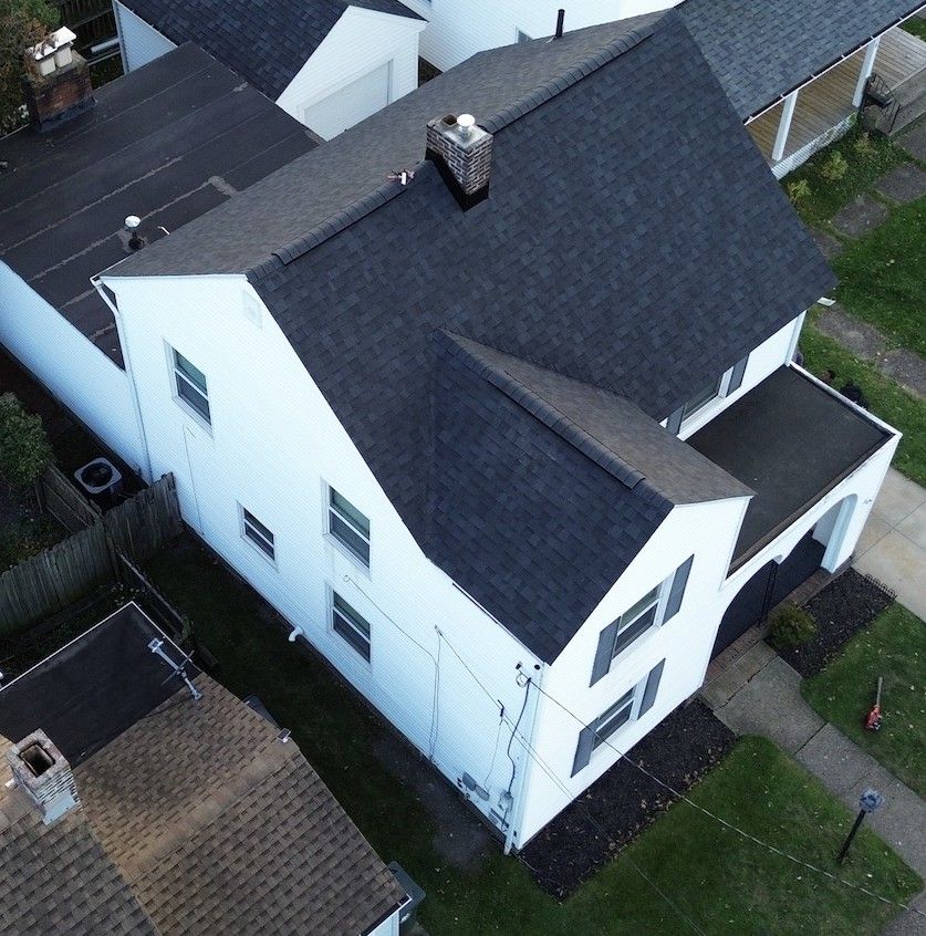 White two-story house with dark roof and trim, chimney, and surrounding greenery.
