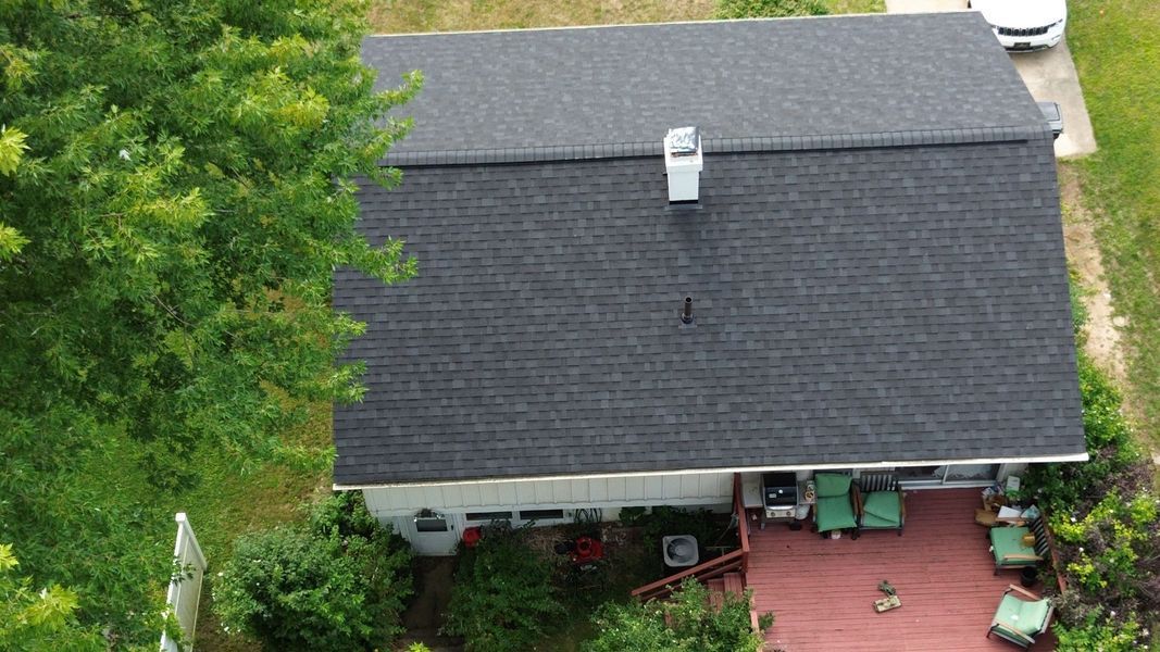 Overhead view of a house with a dark gray shingle roof and a red deck; surrounded by green trees and grass.
