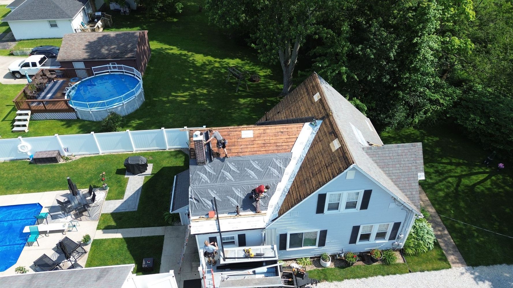 Roofers working on a house with a blue facade, a brown roof, and a pool in the yard.