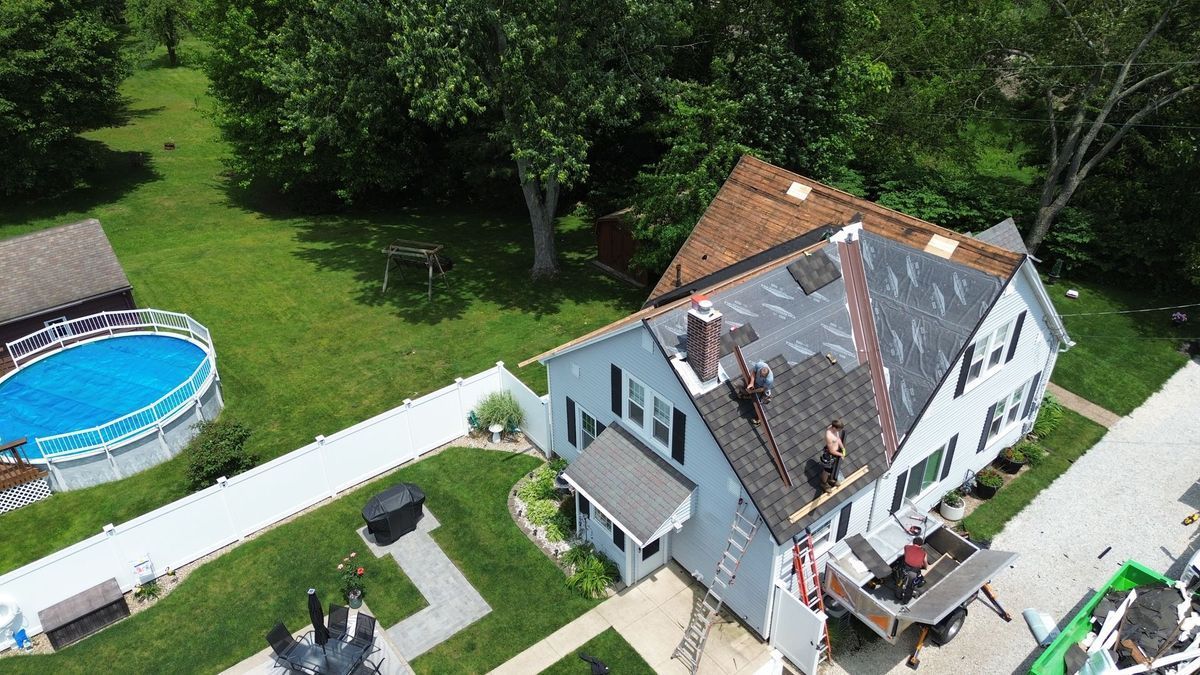 Aerial view of a house with roofers working on the roof; a backyard with a pool and trees is visible.