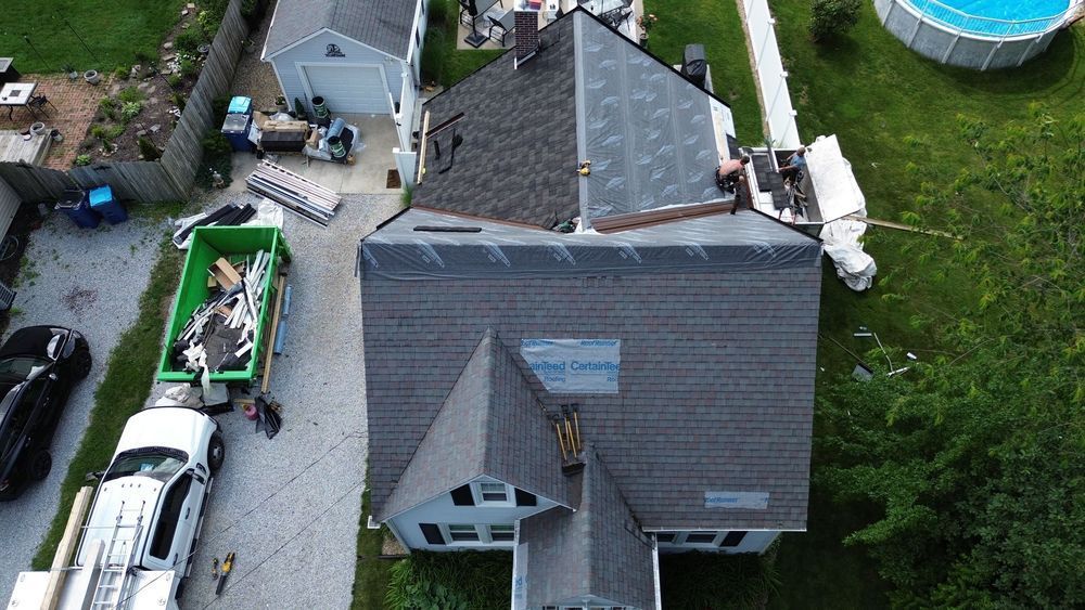 Aerial view of a house with roofing being replaced. Green dumpster and work vehicles are in the driveway.