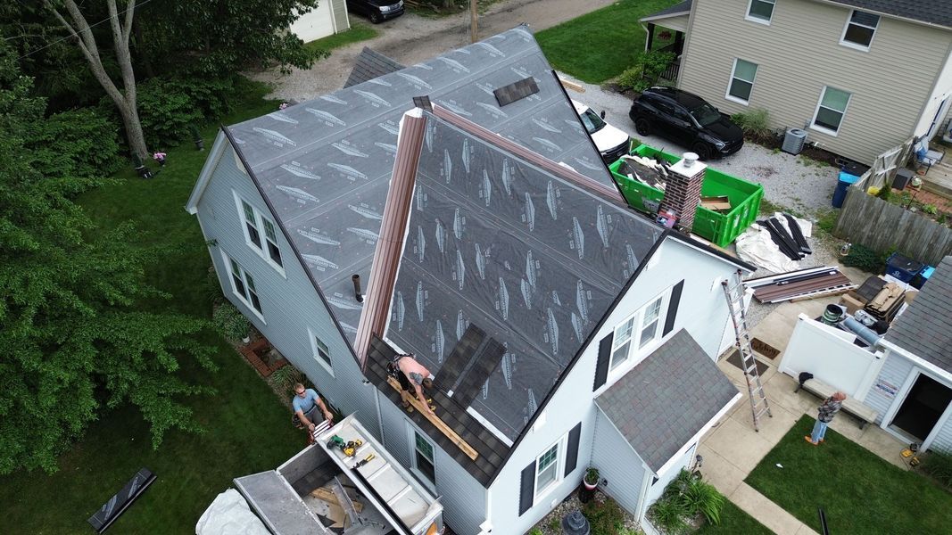 Roofers working on a house roof. Gray siding, black roof shingles, ladder, green dumpster, and foliage.