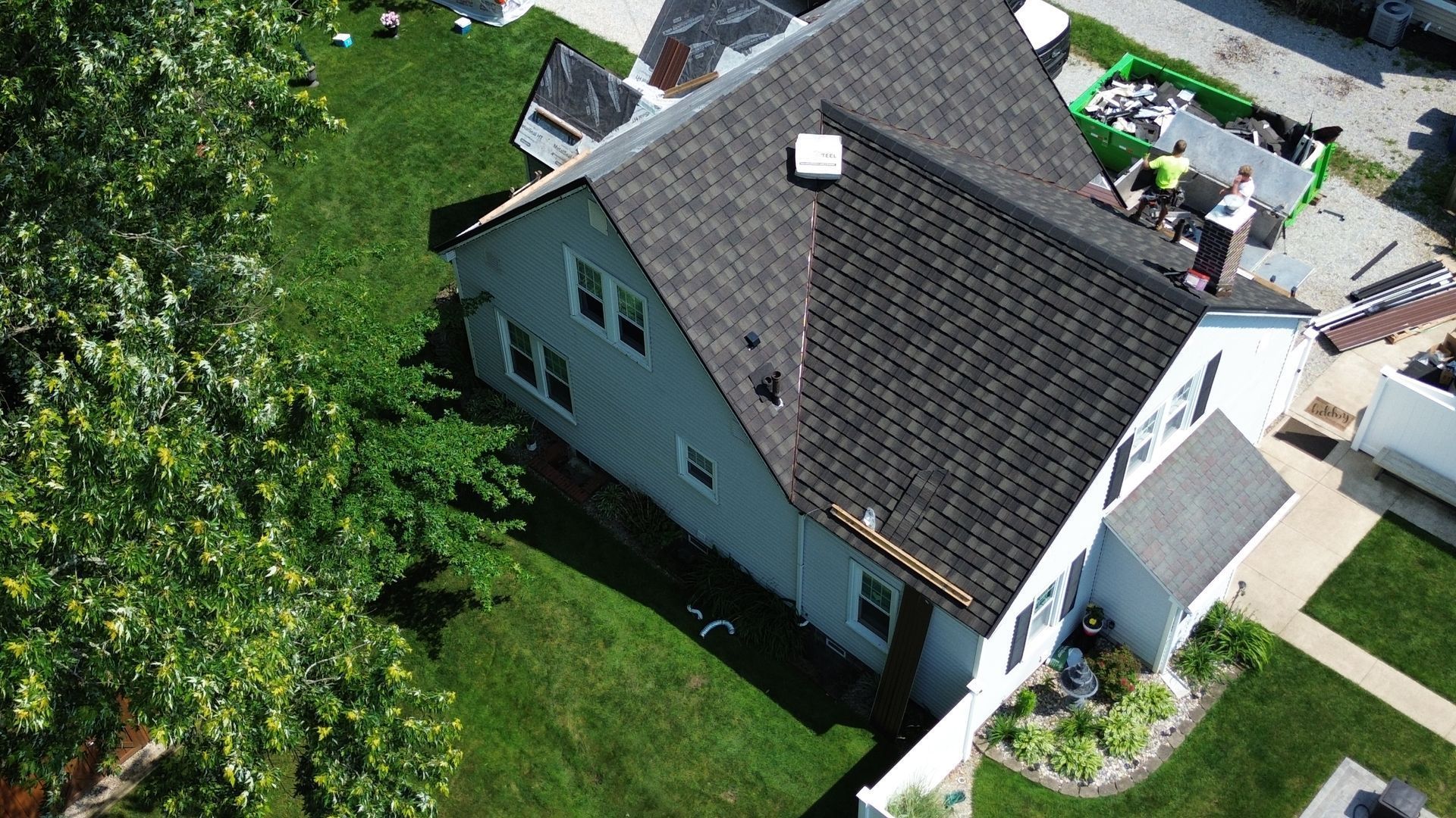 House with roof being replaced; workers on the roof, green grass, a nearby tree.