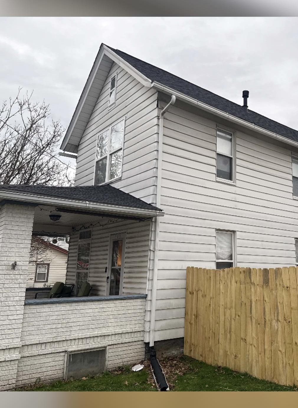Two-story house with white siding and black roof. A wooden fence is along the side of the house.