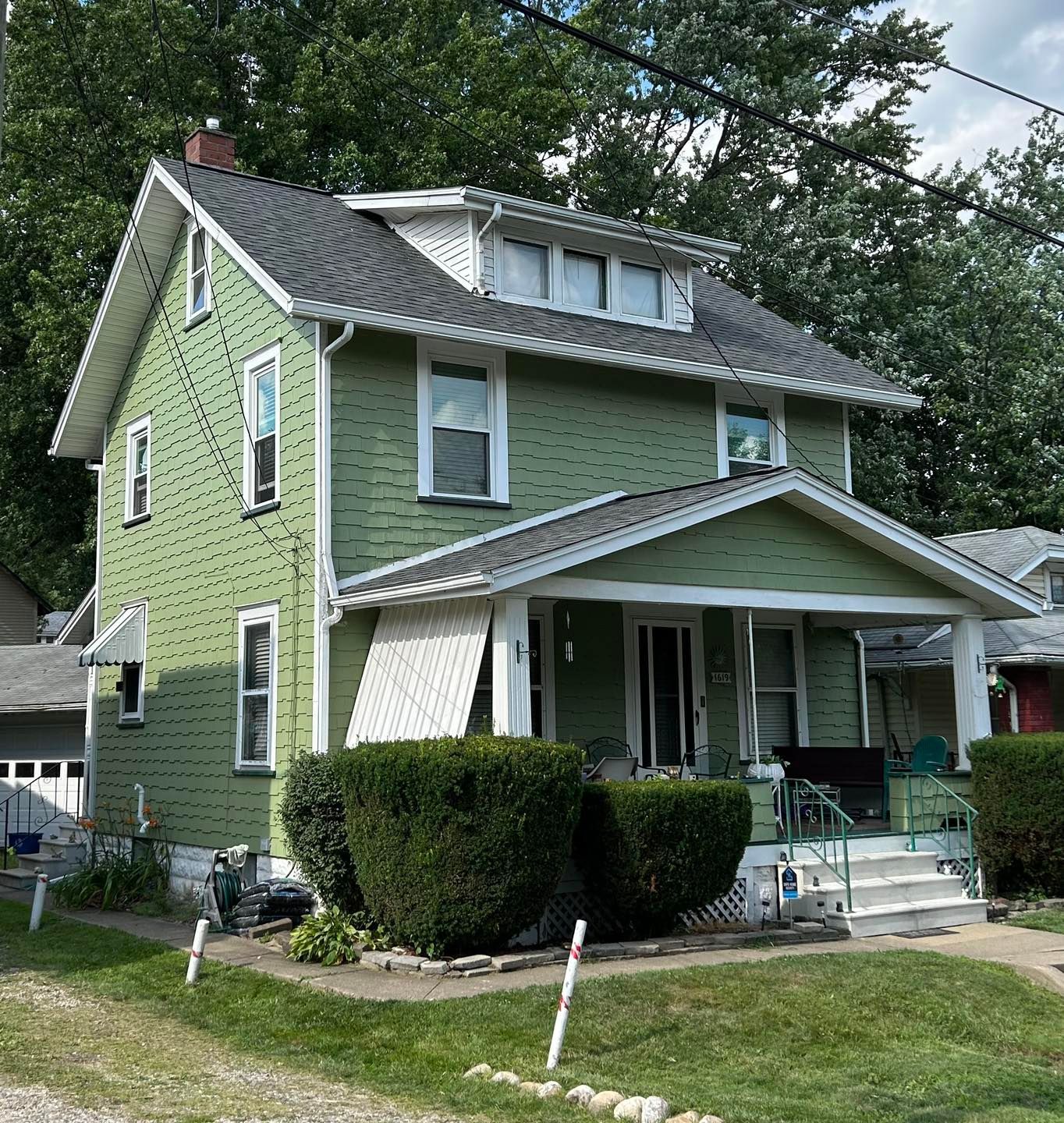 Green two-story house with a porch and bushes in front. The sky is overcast.