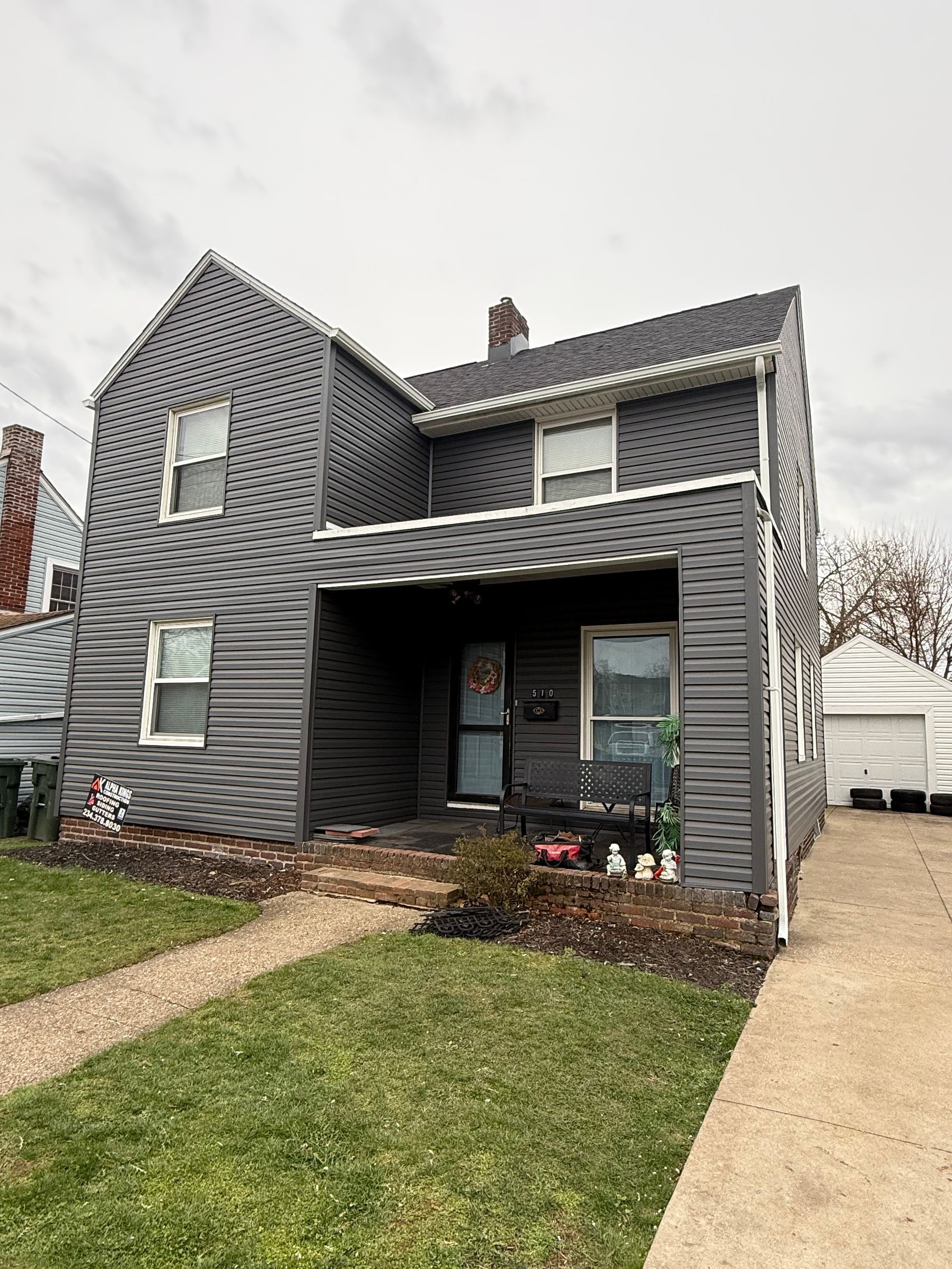 Gray two-story house with front porch, sidewalk, and detached garage on a cloudy day.