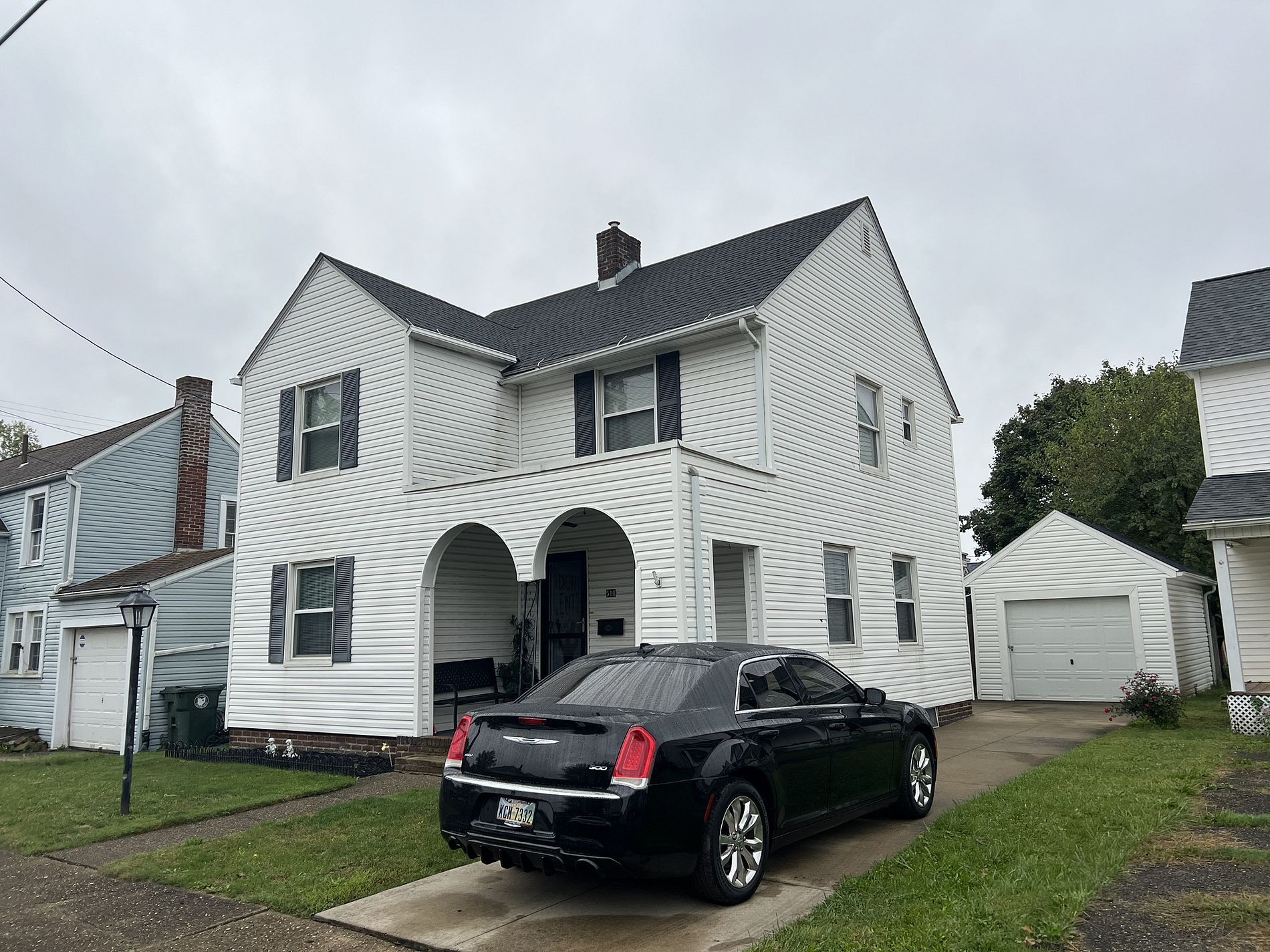White two-story house with black car in driveway. Gray sky. Garage in the background.