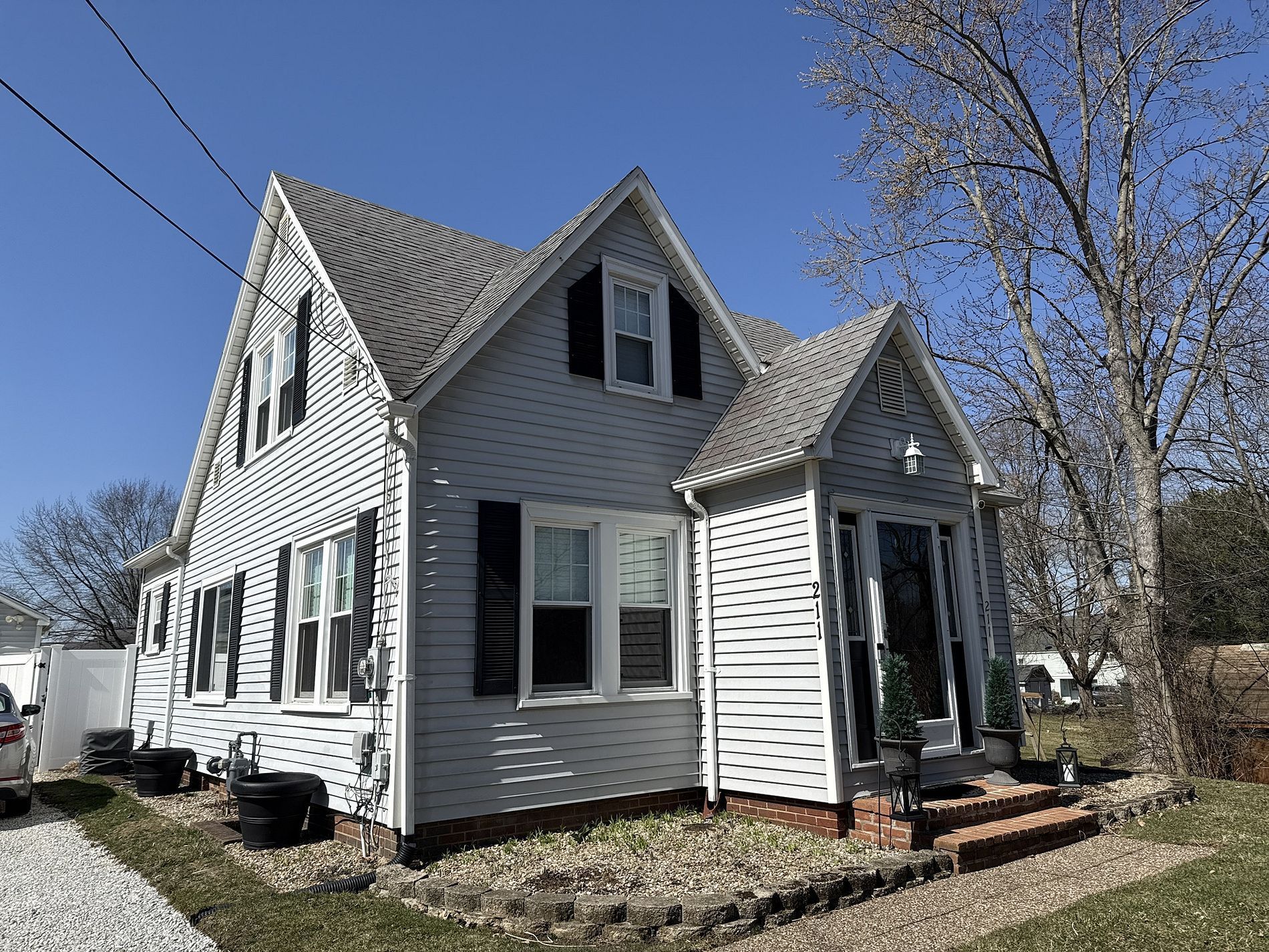Two-story light blue house with black shutters, a gray roof, and a small front yard, under a blue sky.