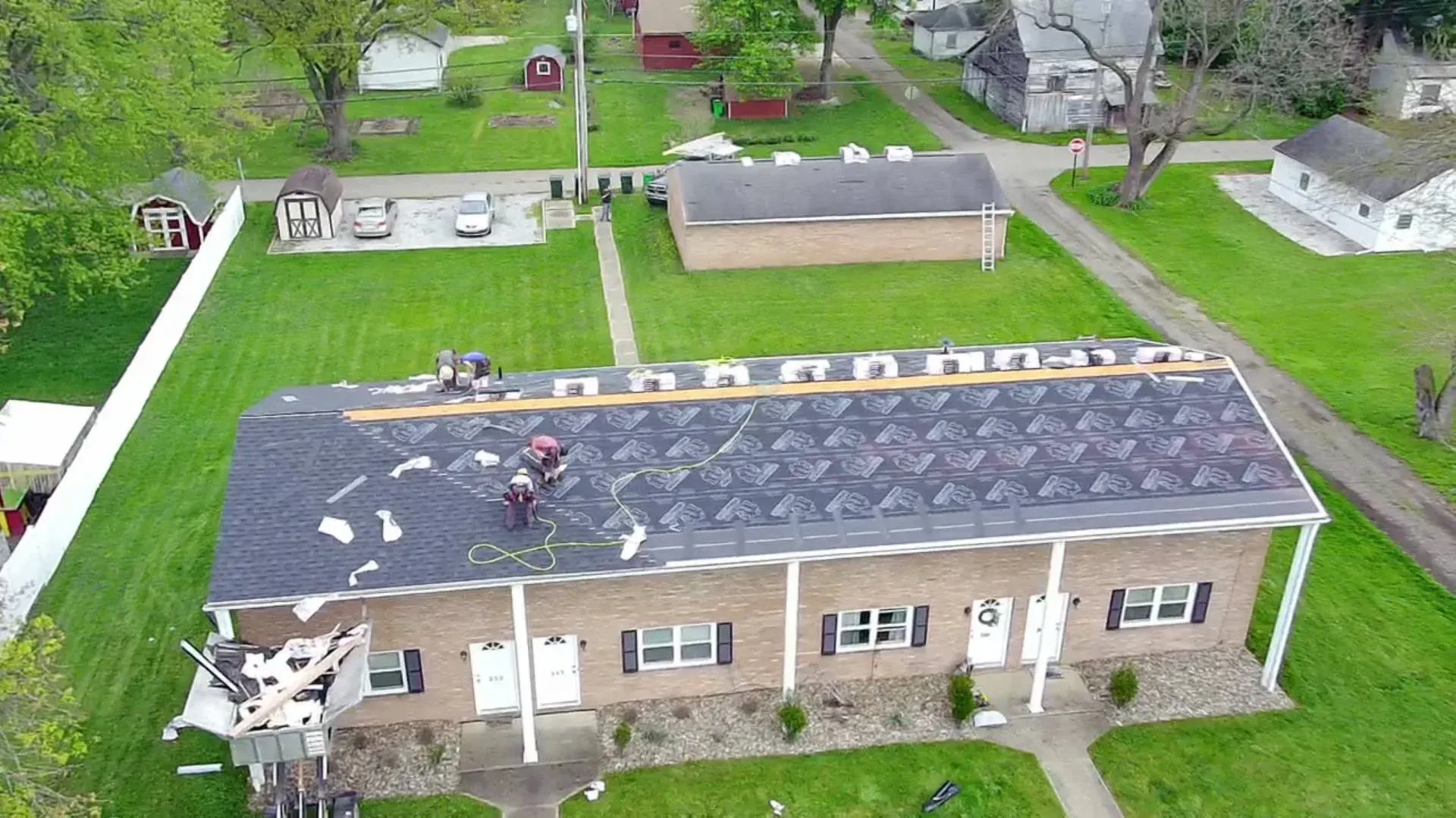 Workers repairing the roof of a brick building. Green lawn, small houses, and a road are in the background.
