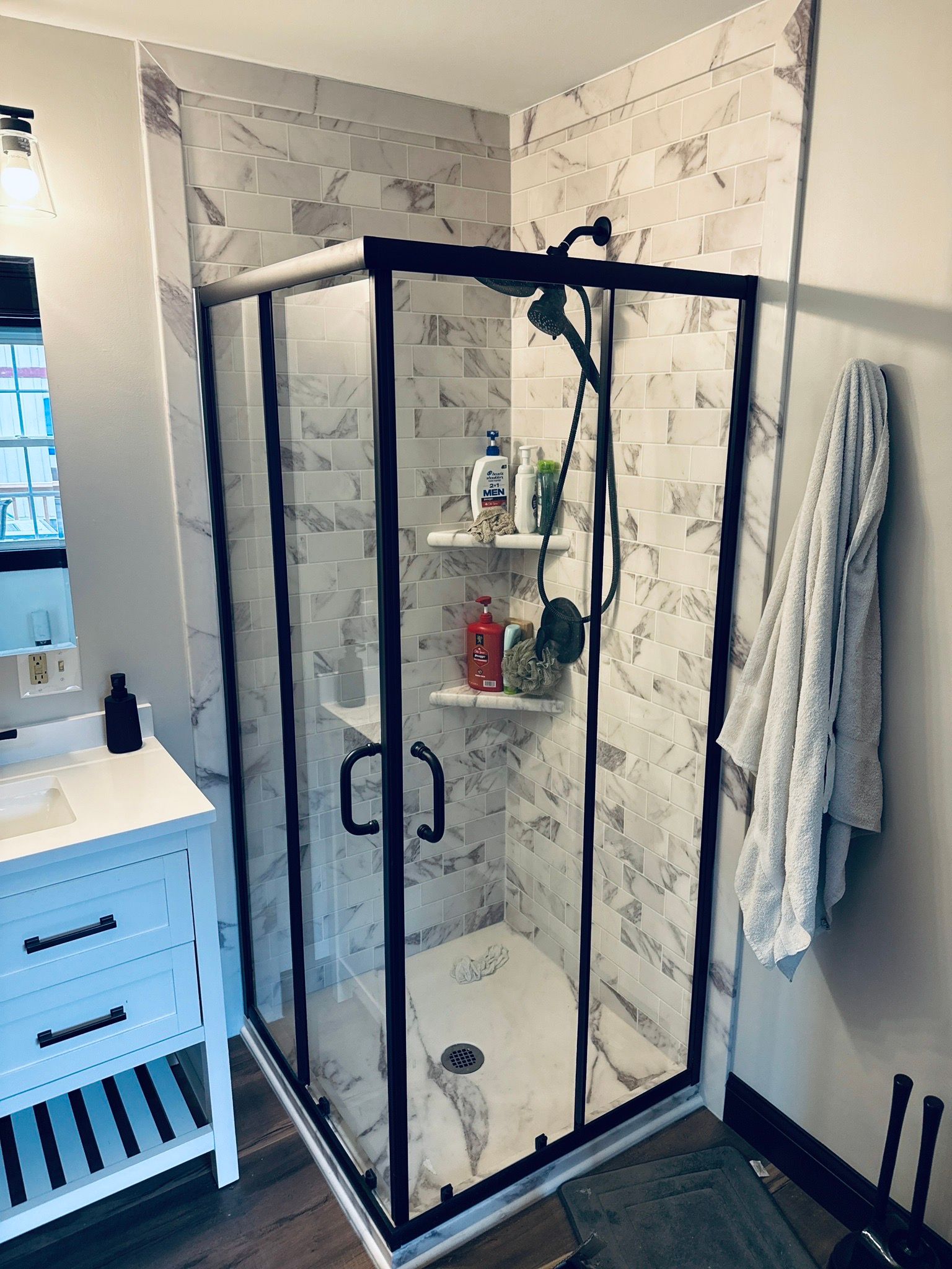 Corner shower with black frame and marble-patterned tile, white shelves, and white vanity.