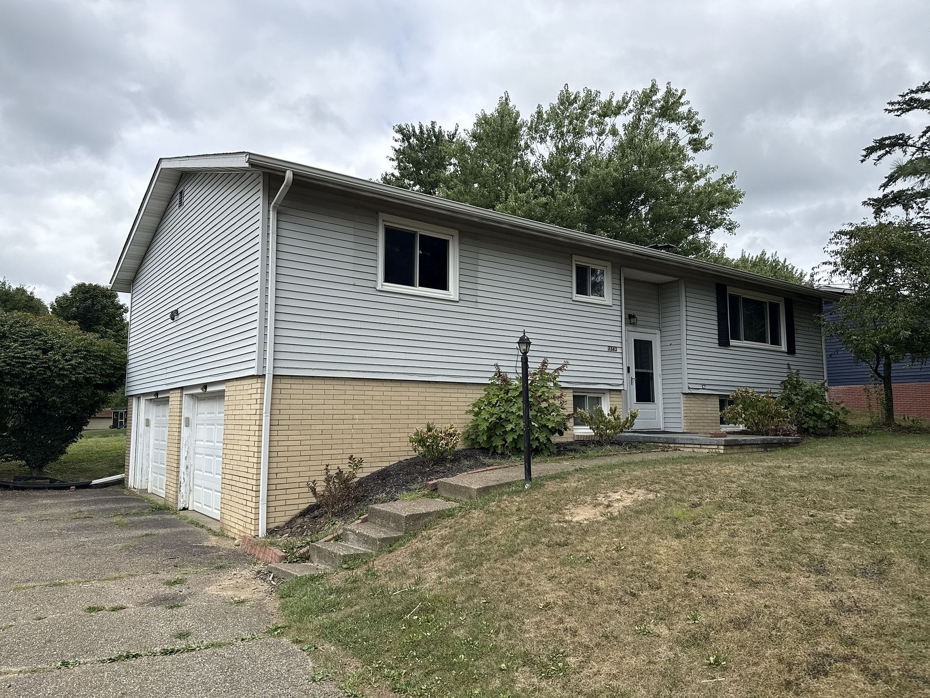 House with light gray siding, tan brick base, driveway, and overgrown lawn under a cloudy sky.