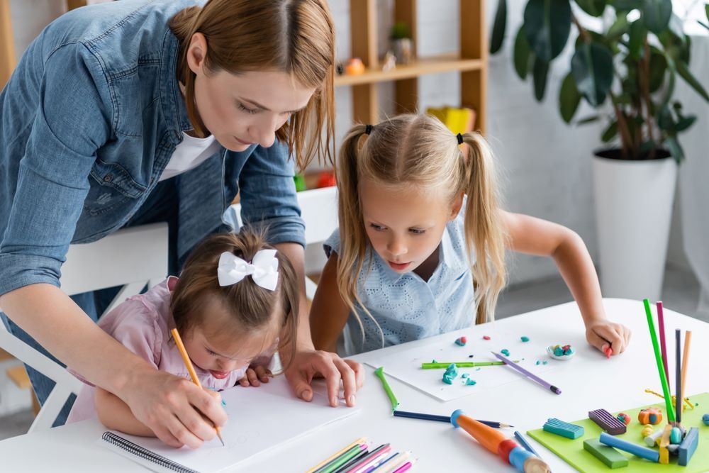 A Woman is Helping Two Little Girls Draw at a Table — Grandma Rosie’s Quality Long Day Care In Horsley, NSW