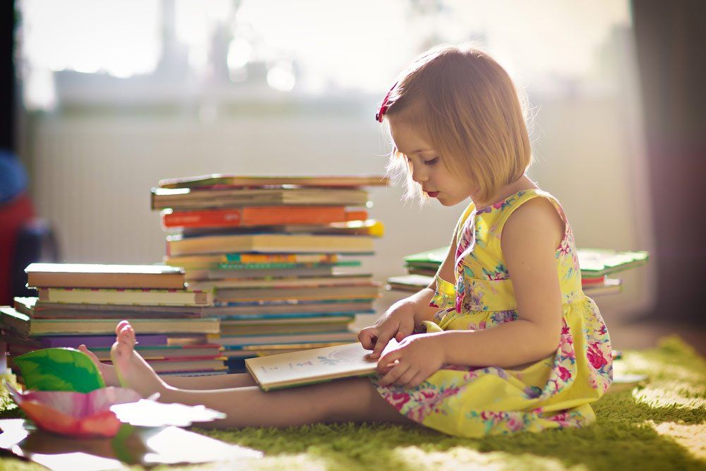 A Little Cute Girl In A Yellow Dress Reading A Book Sitting On The Floor — Grandma Rosie’s Quality Long Day Care In Wollongong, NSW
