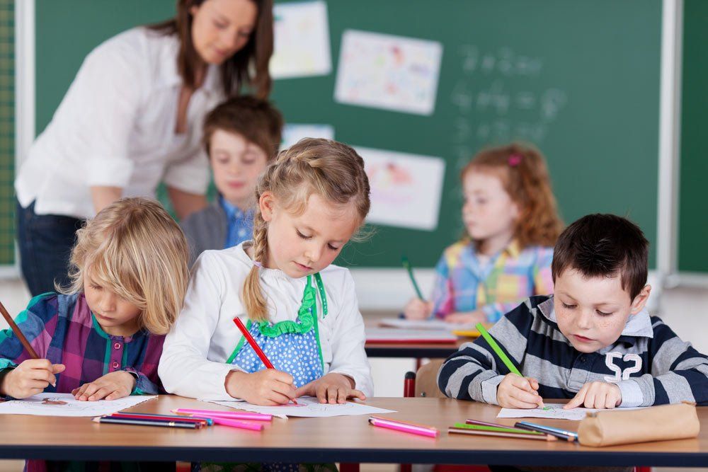 Two Girls Drawing With Pencil Crayons At The Front As The Teacher Works With Pupils At The Back — Grandma Rosie’s Quality Long Day Care In Wollongong, NSW