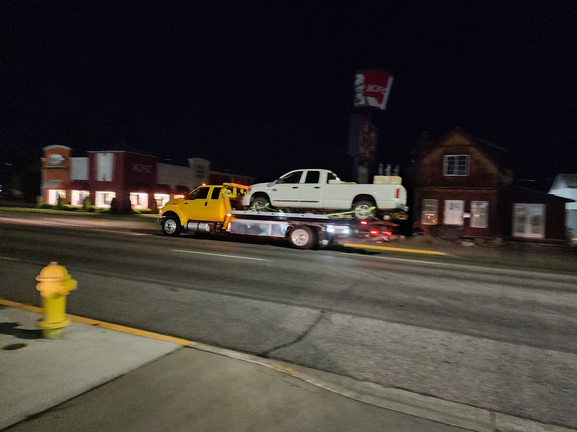 A yellow tow truck is towing a white truck down a street at night.