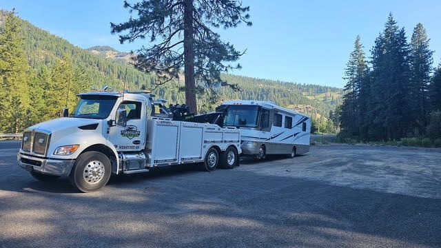 A white tow truck is towing a rv in a parking lot.