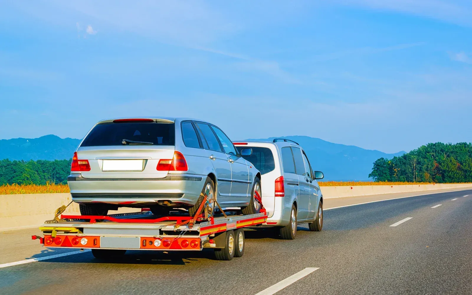 Car being towed on a flatbed trailer on a highway. Clear blue sky and green hills in the background.