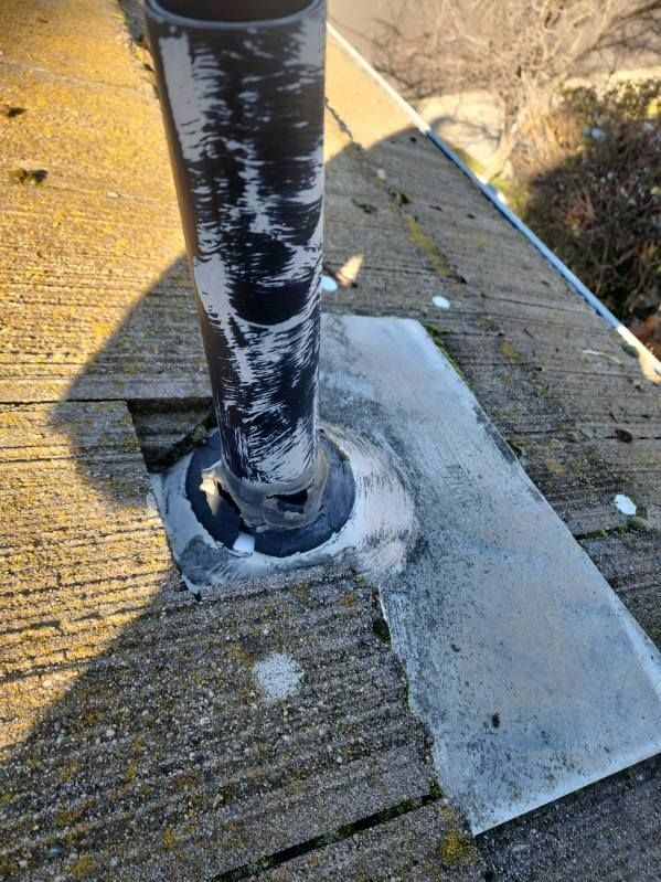 Black pipe on a weathered wood surface.  A metal flashing surrounds the pipe and sealed with gray sealant.