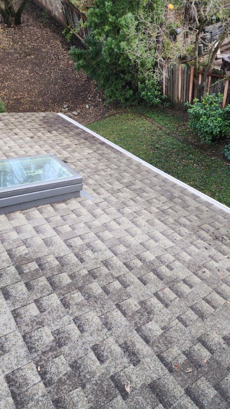 Shingled roof with a skylight, against a backdrop of a grassy hillside and wooden fence.