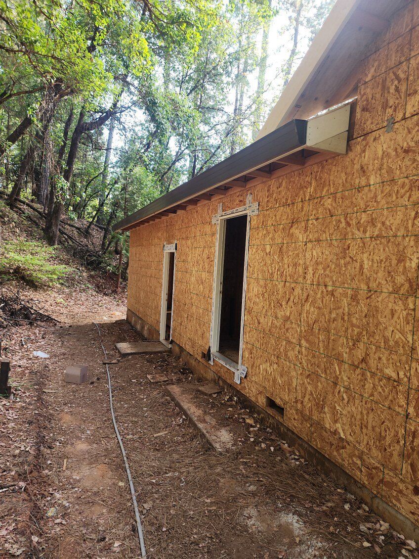 Building exterior with OSB sheathing, door/window openings, and a brown roof in a wooded area.