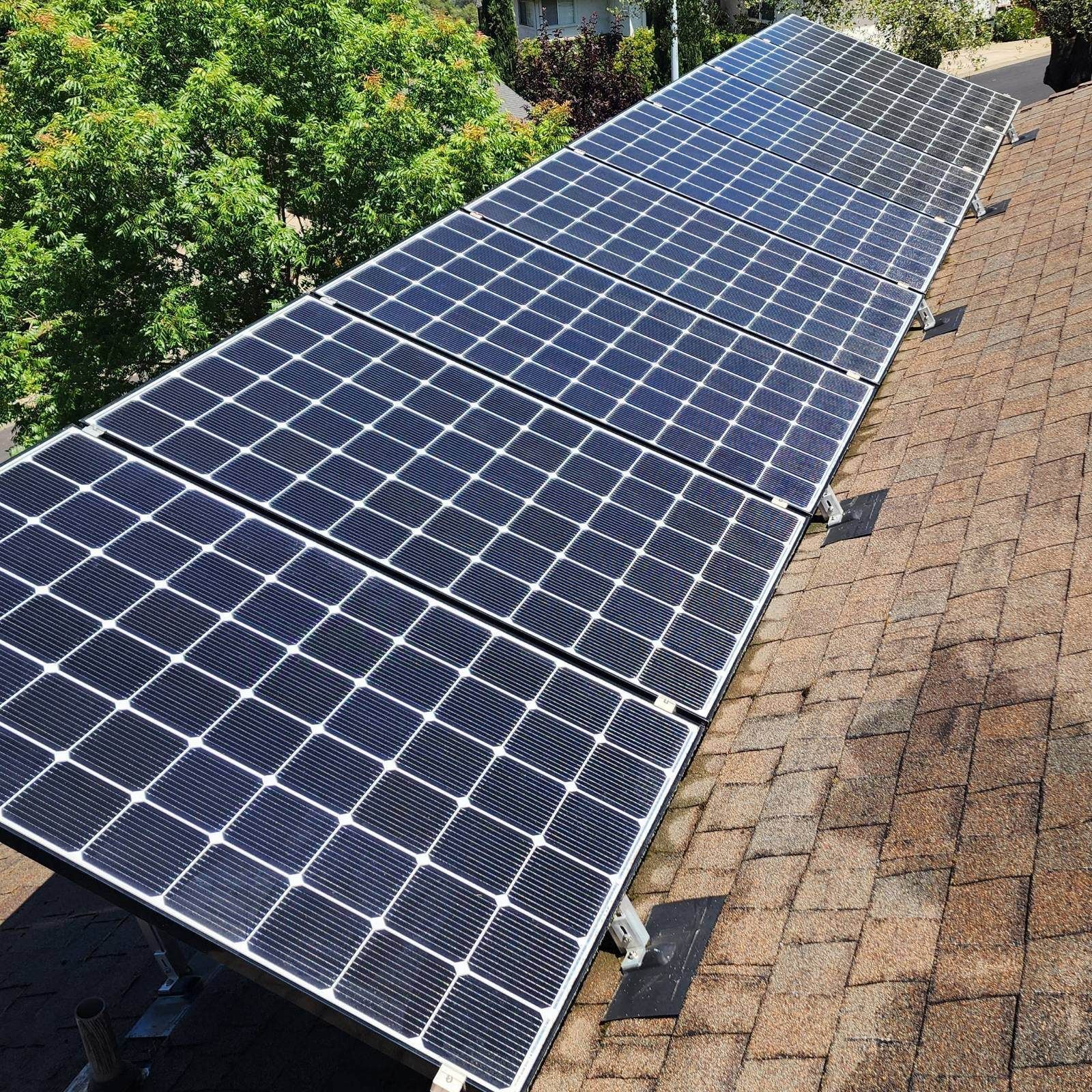 Solar panels installed on a shingled roof, reflecting sunlight. Green trees are visible in the background.