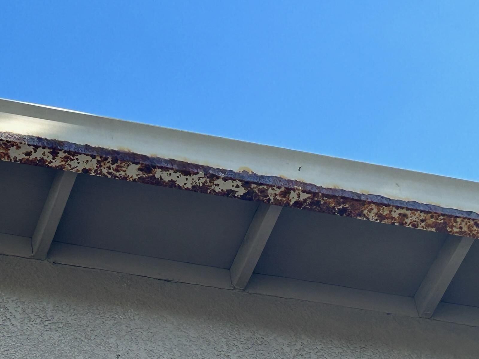 Rusted metal gutter against a blue sky, part of a white building's overhang.