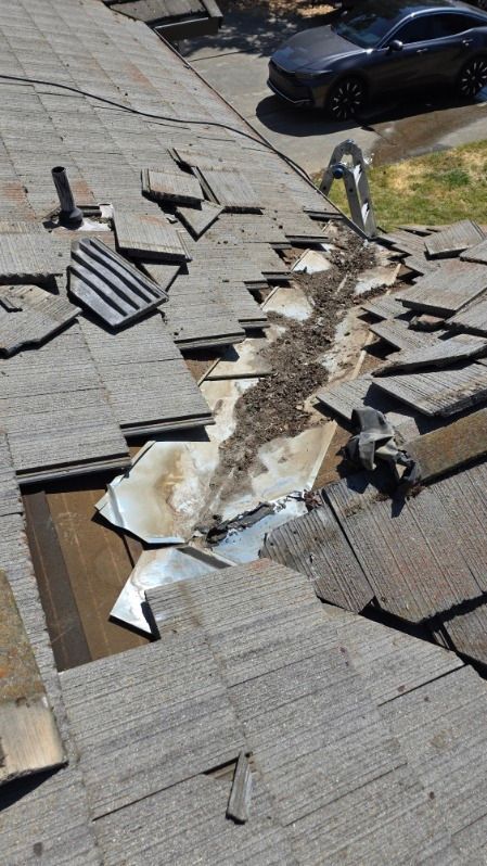 Damaged asphalt shingle roof with missing sections, debris, and a metallic flashing. A car is parked nearby.
