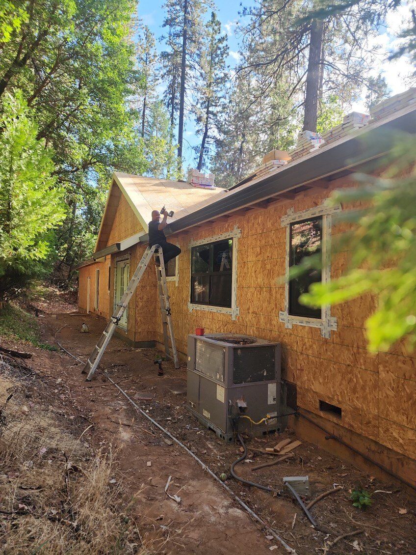 Construction worker on a ladder, near a partially built house with exposed wood siding. Green trees and blue sky.