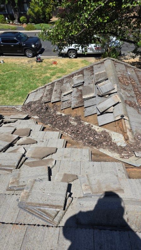 Damaged roof with missing and loose shingles, debris visible. Shadow of person taking the photo.