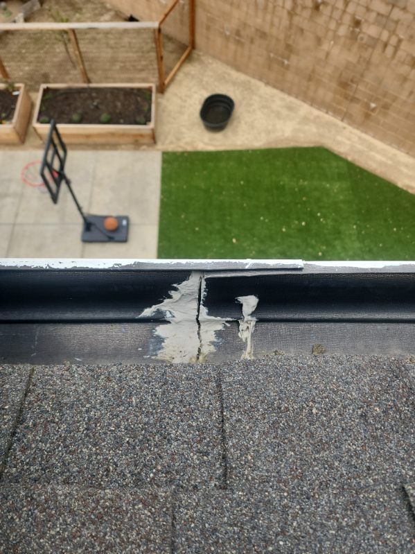 Close-up of roof with asphalt shingles and a damaged gutter filled with grey sealant. Backyard in background.