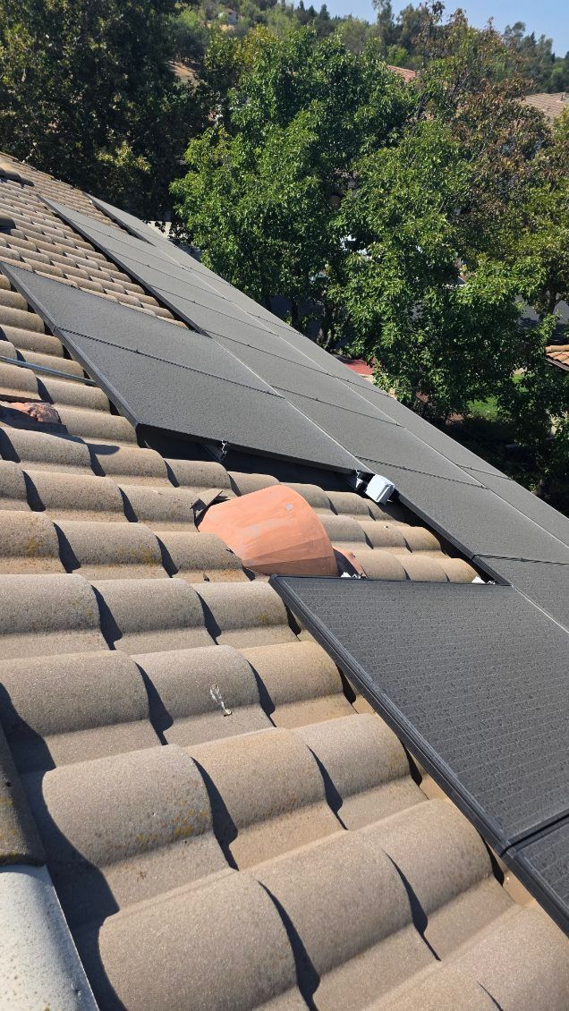 Solar panels installed on a tiled roof. Some tiles are missing. Trees and sky in the background.