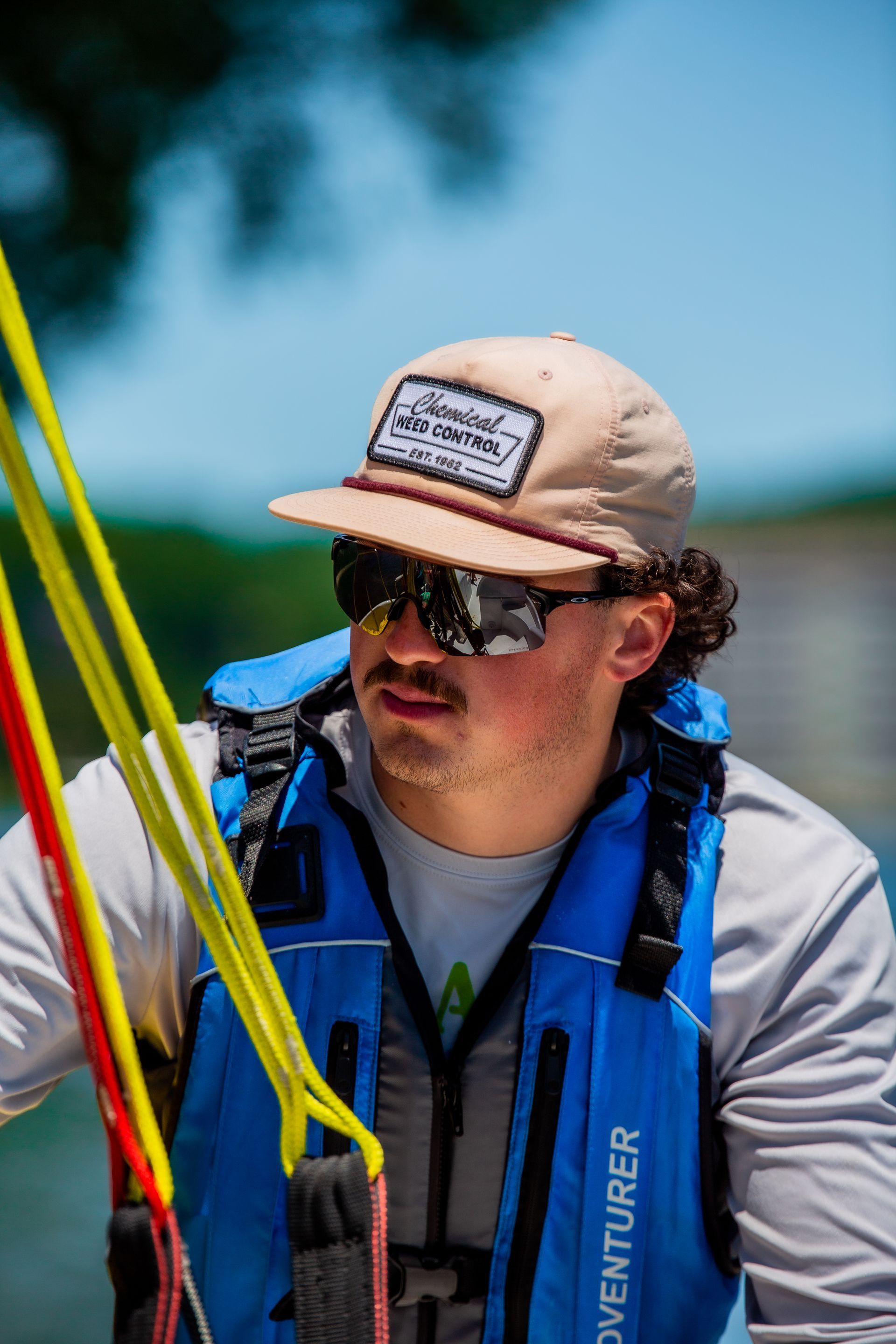 Man wearing sunglasses, hat, and life vest, looking off-camera. Holding ropes with yellow and red stripes; blue sky in background.