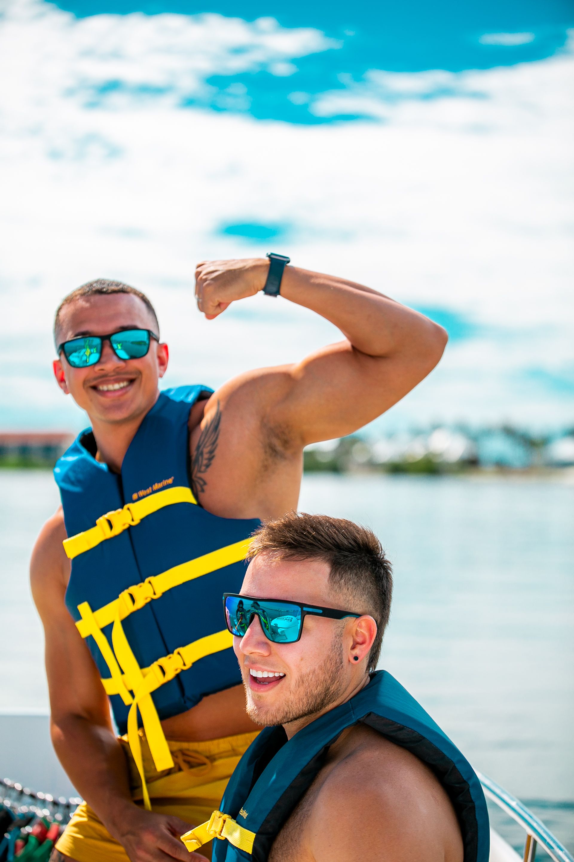 Two men on a boat, wearing sunglasses and life vests. One flexes a bicep, smiling; the other smiles. Blue sky backdrop.