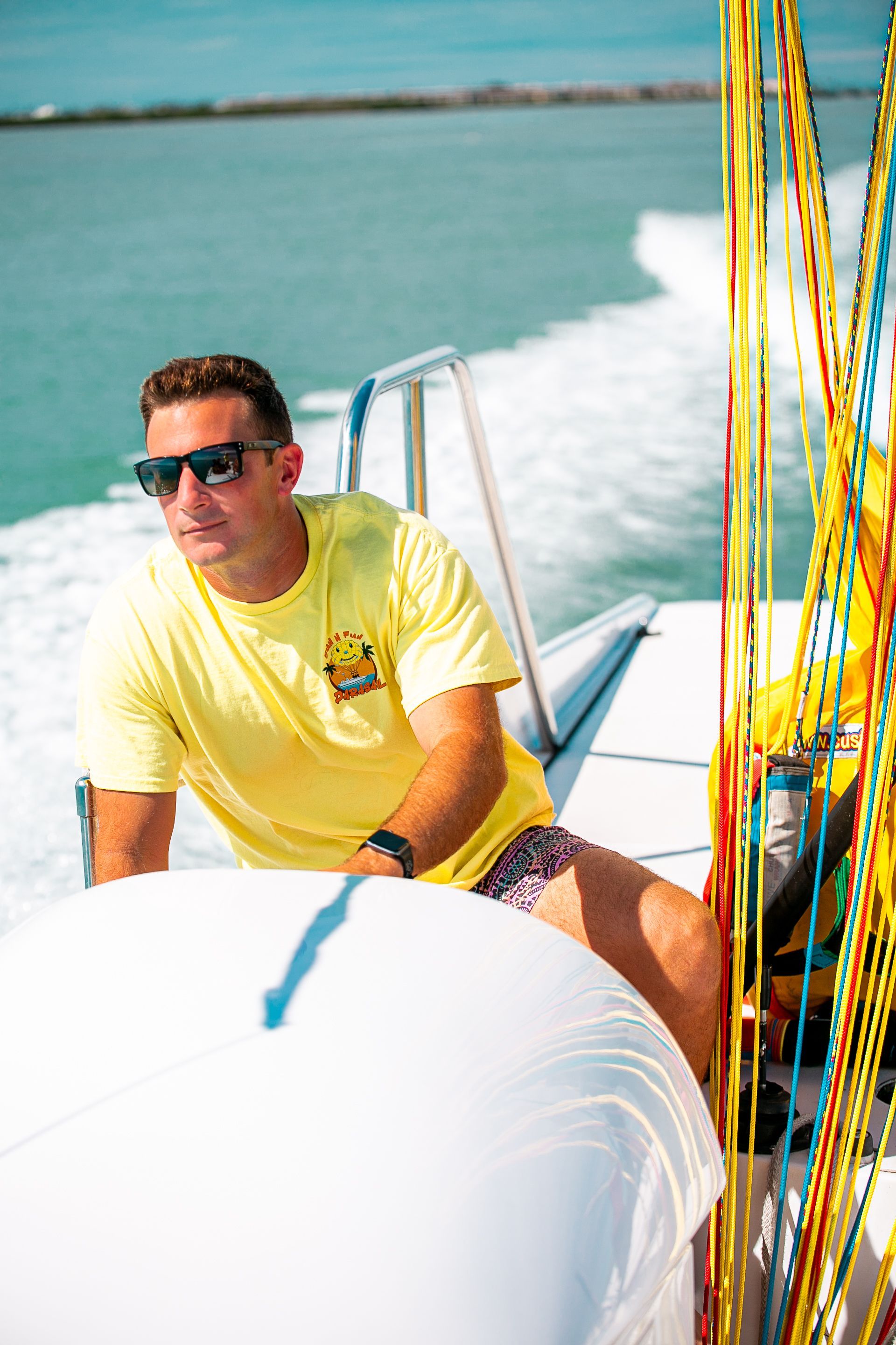 Man in sunglasses driving a boat, yellow shirt, pulling parasail lines on sunny water.