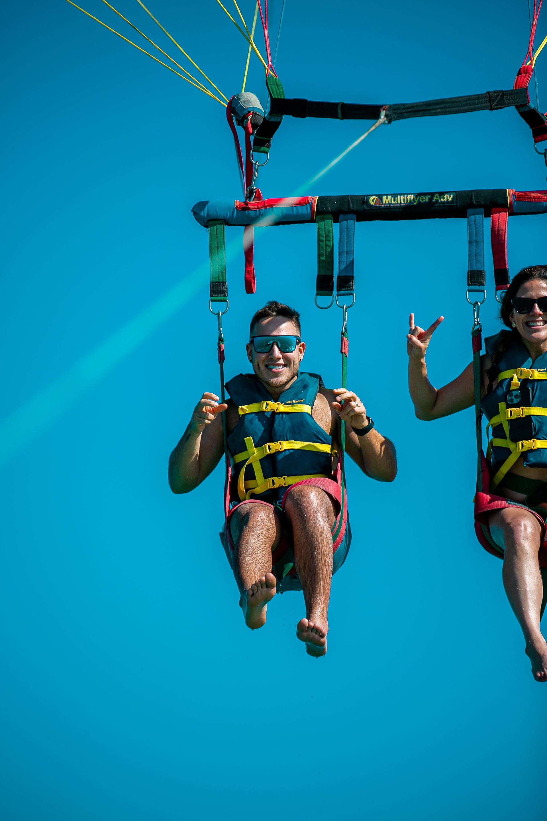 Two people parasailing against a bright blue sky, smiling and wearing life vests.