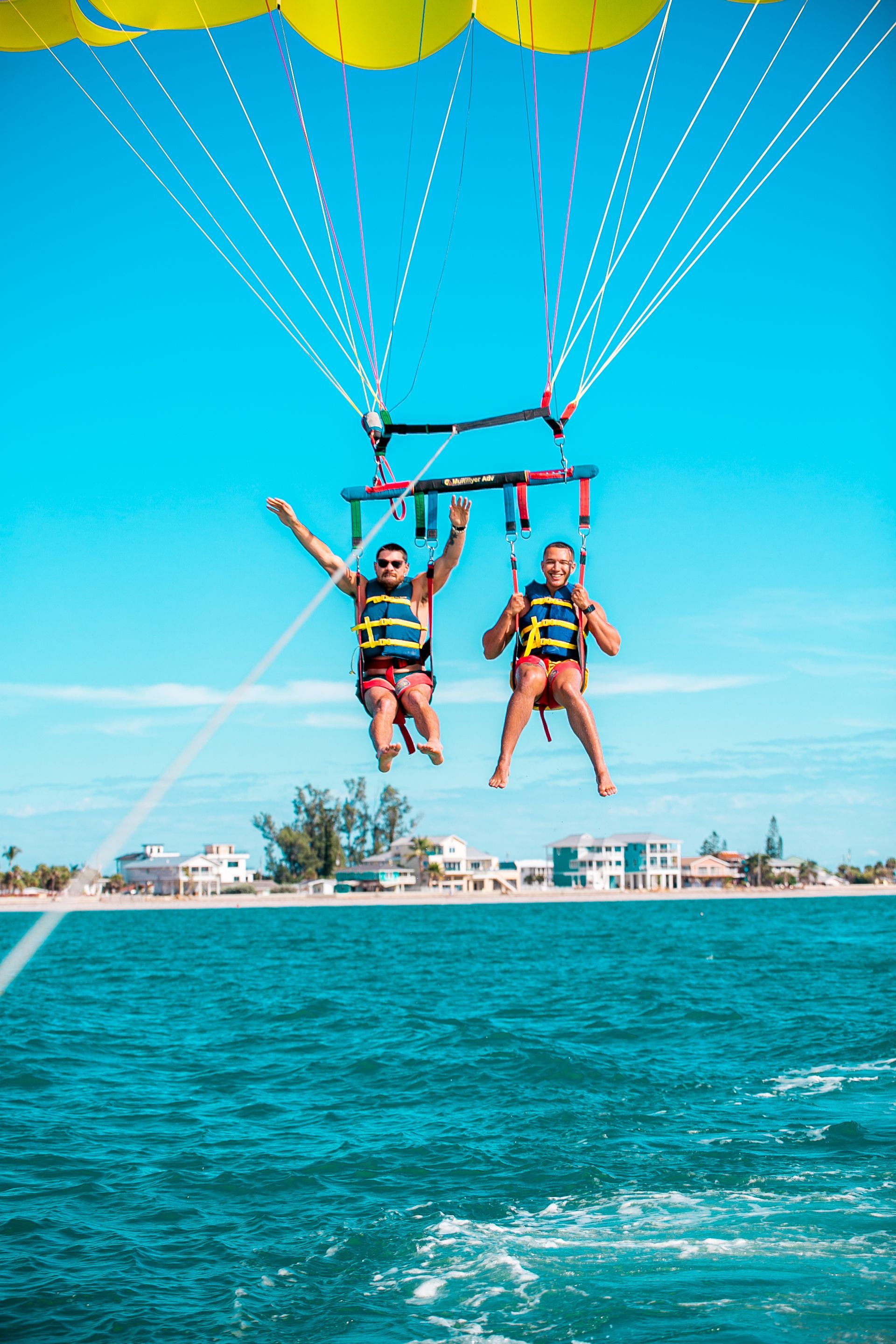 Two people parasailing over ocean with blue sky and shoreline in the background.