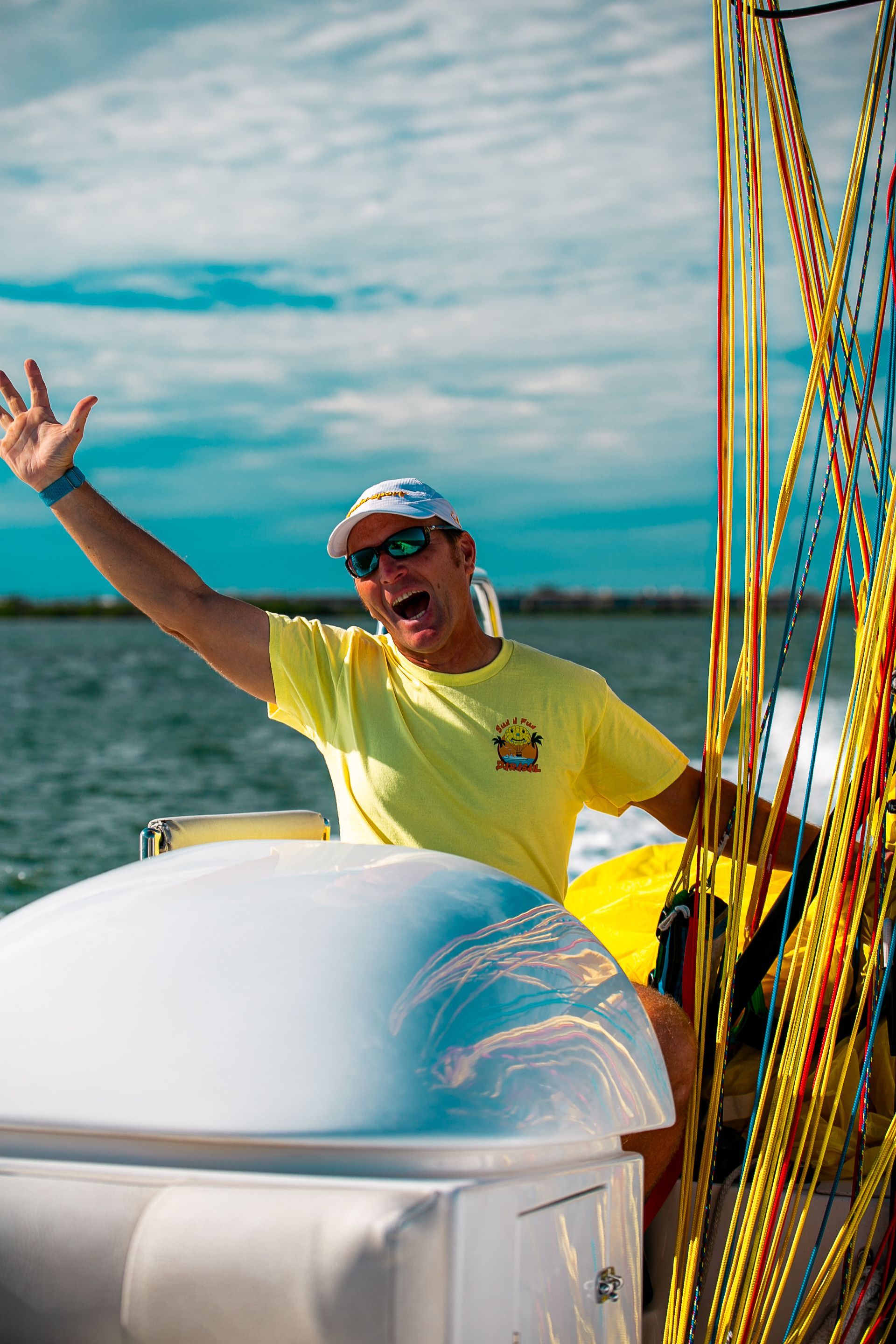 Man on boat with arm raised, smiling. Yellow shirt, sunglasses, blue sky, water.