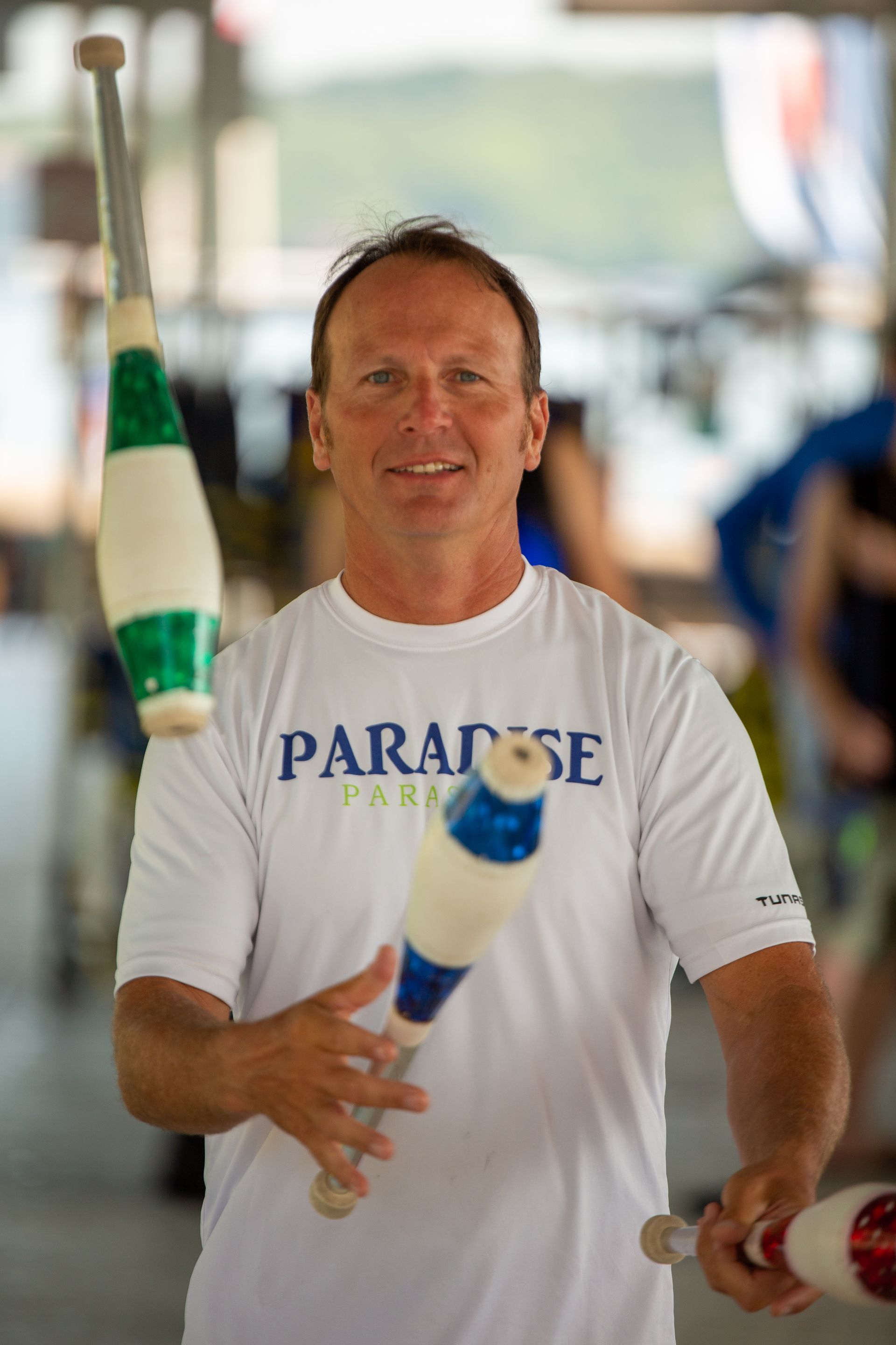 Man juggling three clubs outdoors. Wearing a white shirt that says 